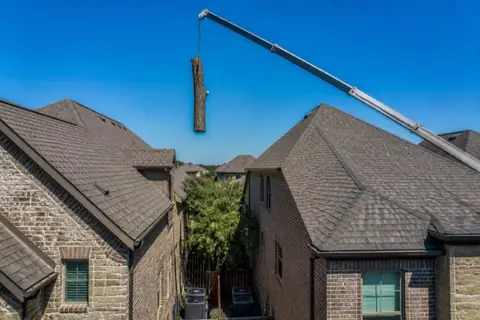 Crane boom reaching over a two-story brick home in a dense subdivision with a whole oak trunk section suspended above the ridgeline