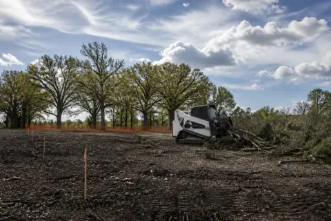 Mini skid-steer with a grapple stacking cedar and scrub on rural acreage while a flagged post oak grove stands preserved behind orange fencing