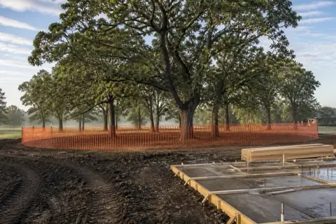 Mature post oak grove ringed by orange protection fencing at the dripline on a new-construction lot with formed slab and stacked lumber behind